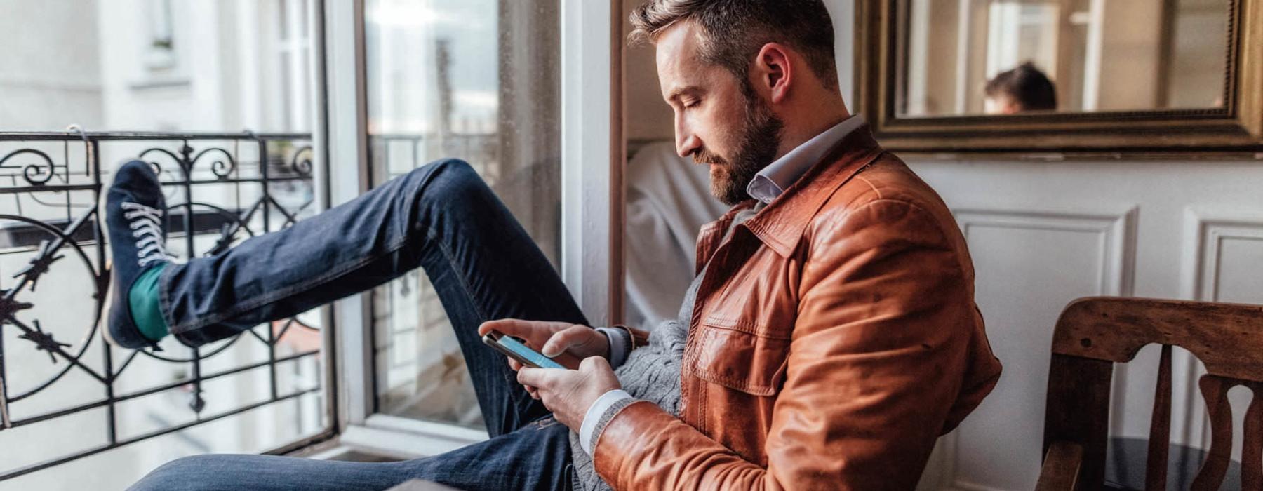 a man sits on a chair and rests his foot on a balcony railing as he looks at his phone in his room